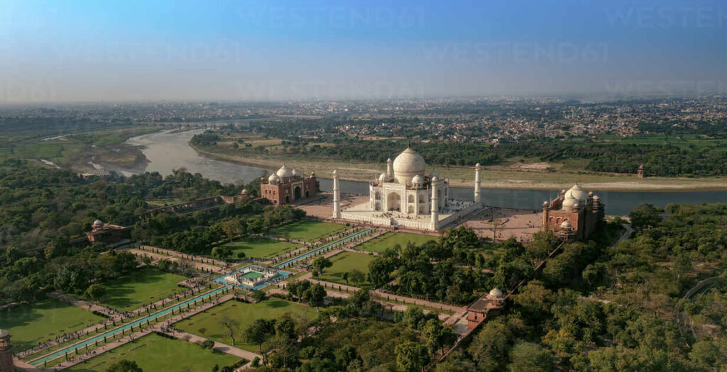 Aerial view of the Taj Mahal complex, gardens, and Yamuna River with the Agra cityscape in the background.